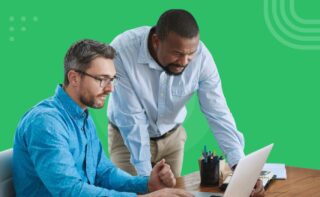 Two men collaborating at a desk, looking at a laptop screen against a green background