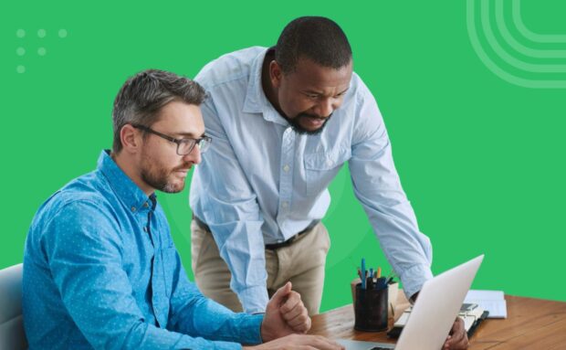Two men collaborating at a desk, looking at a laptop screen against a green background