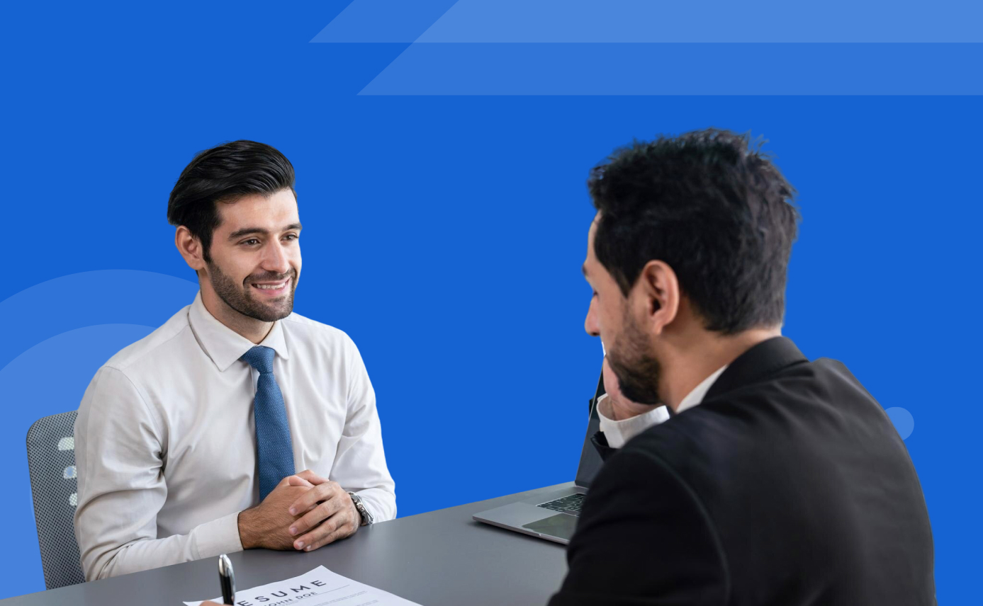 Two men in business attire sit across a desk during a job interview, with a resume and laptop visible.