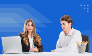 A woman and a man sitting at a desk looking at her laptop in front of a royal blue background.