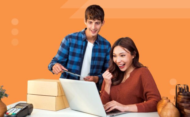 Two people excitedly reviewing content on a laptop near shipping boxes on an orange background