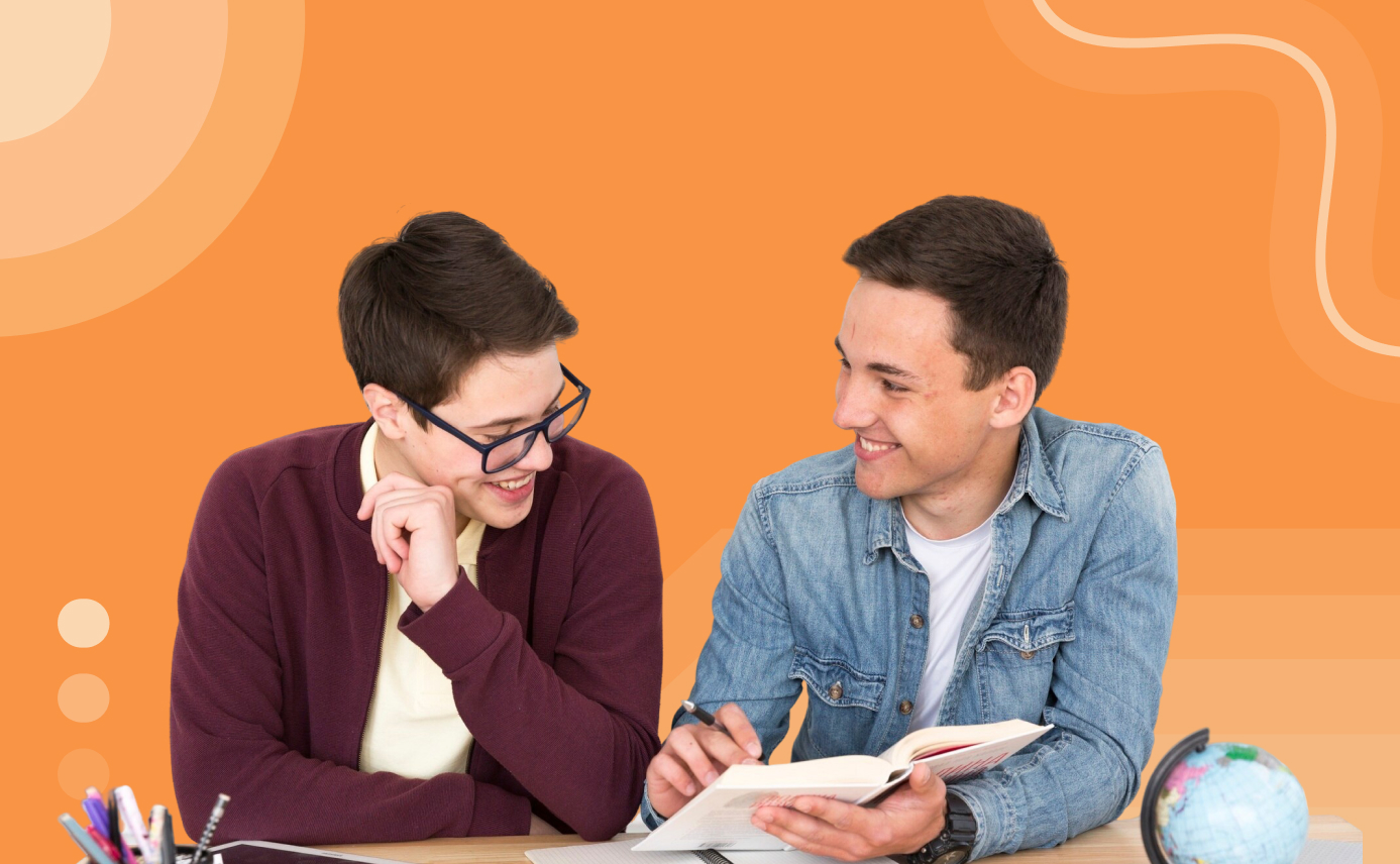 Two me sitting next to each other at a desk smiling and one of the men is pointing something out in a book for the other.